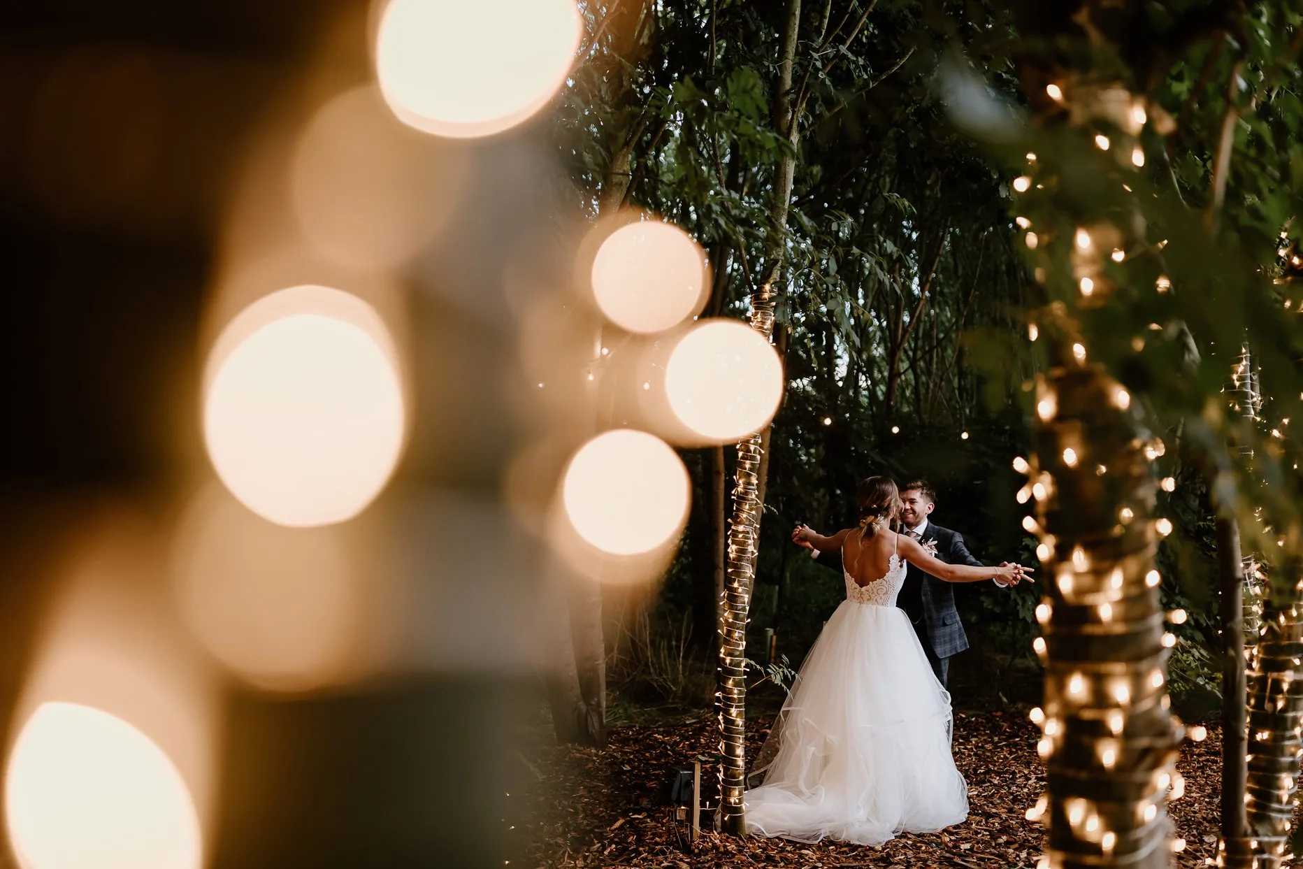Bride and groom dancing in between the trees on the woodland dancefloor at Oaklands. The trees are decorated with gold fairylights.