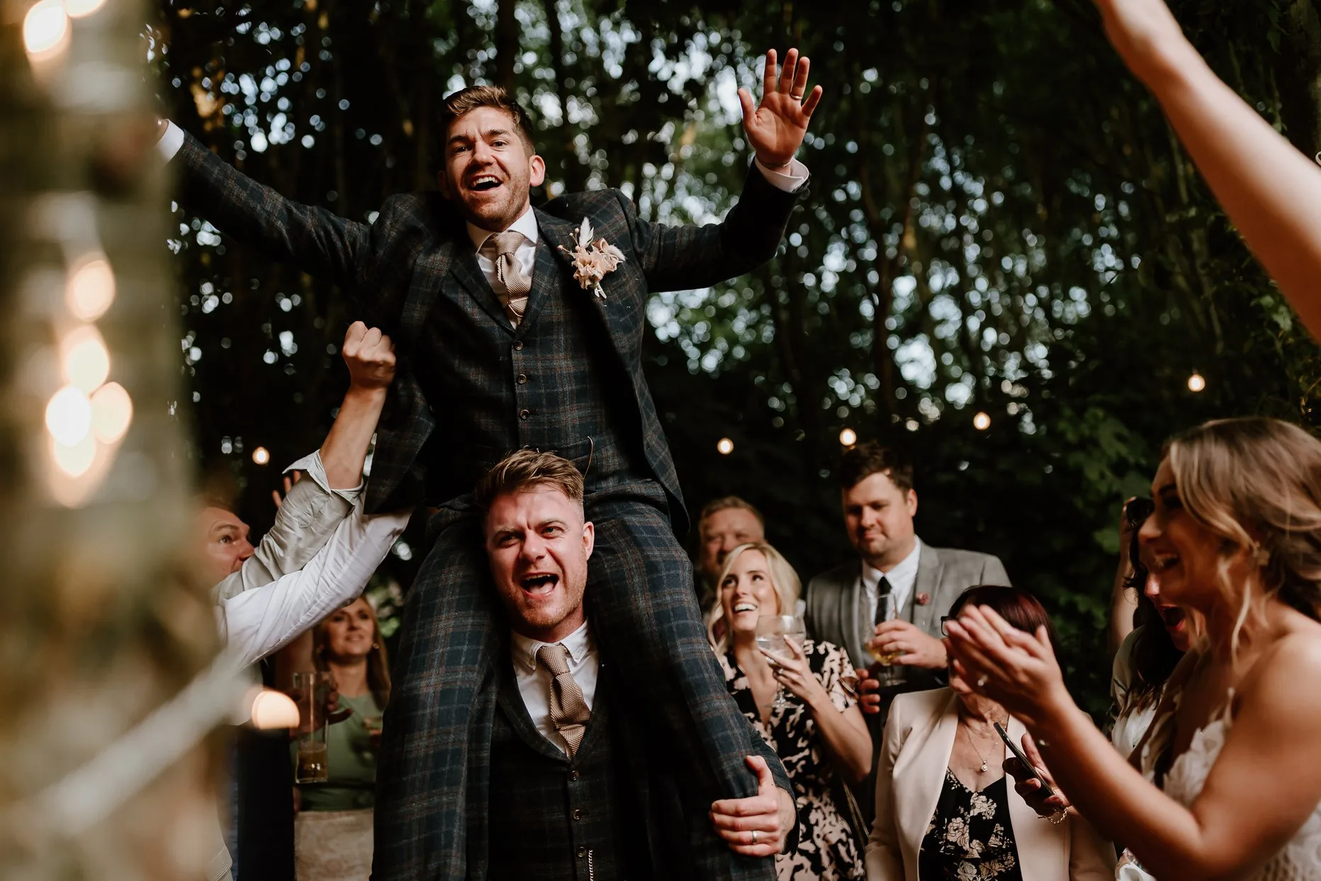 Groom on top of a groomsman shoulders dancing in the woods at Oaklands Grand Lodge wedding venue in Driffield. Wedding guests surround them clapping and smiling.