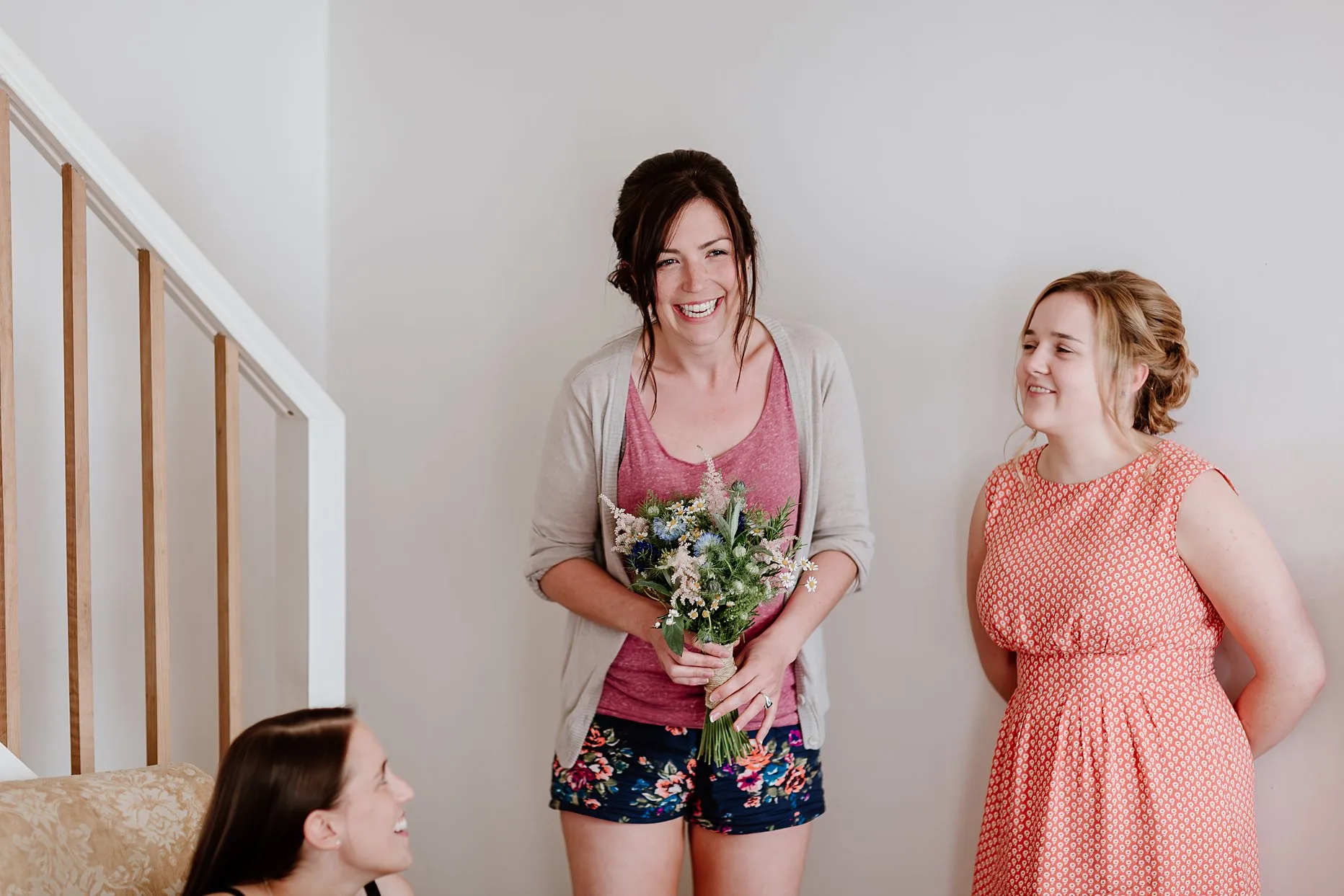 Bride holding wedding bouquet and smiling. Her bridesmaids are stood next to her all dressed in casual clothing. 