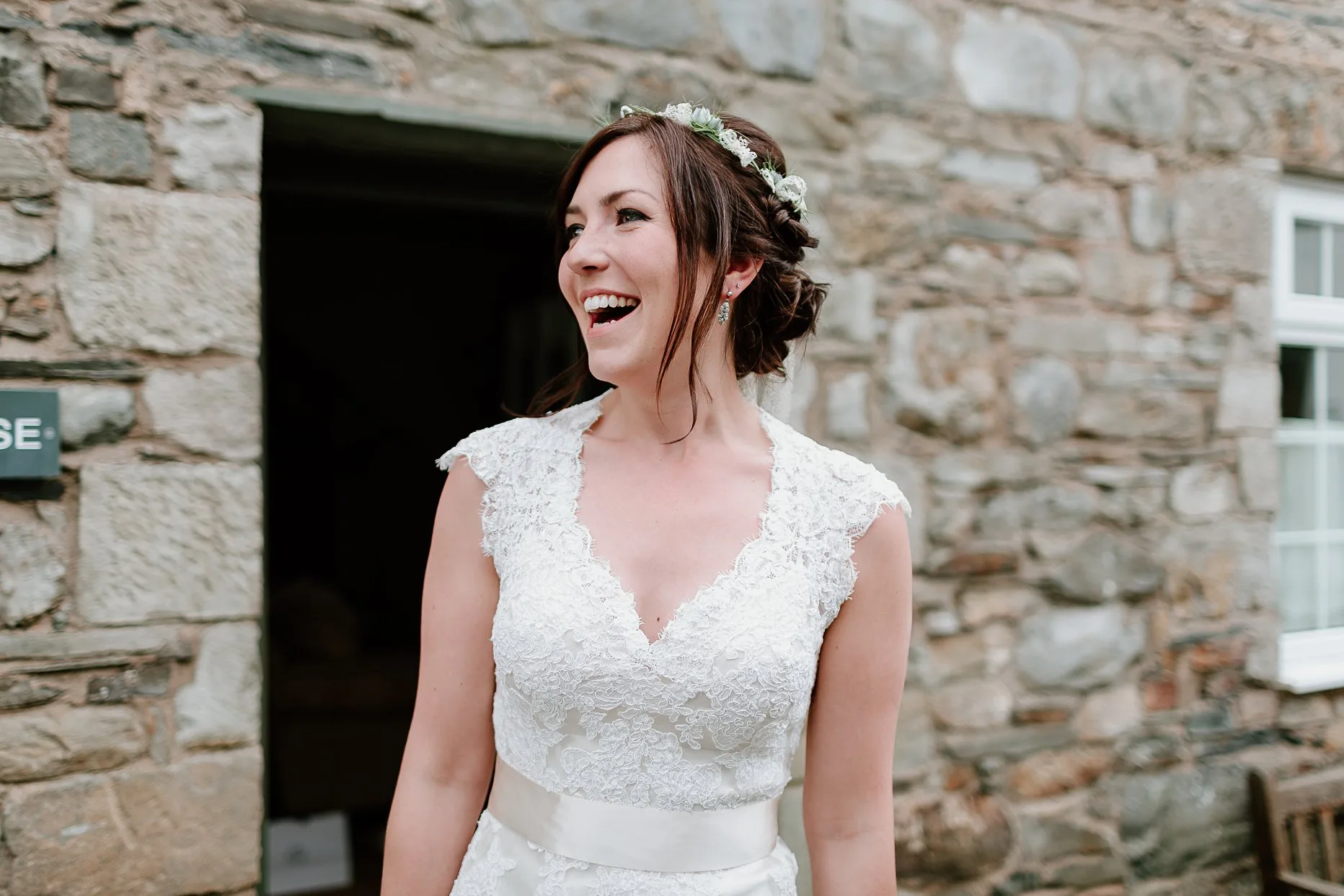 Bride dressed in boho lace wedding dress and flower crown stood outside of a stone bricked building. Bride is  laughing at something in the distance.