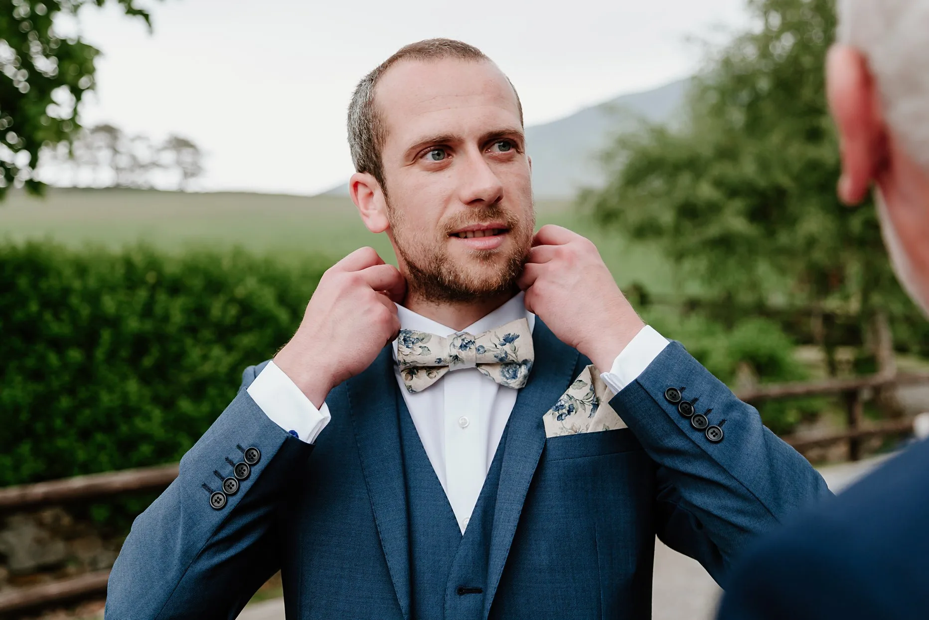 Groom dressed in blue suit ad floral bowtie. Groom is adjusting his collar ad looking into the distace.