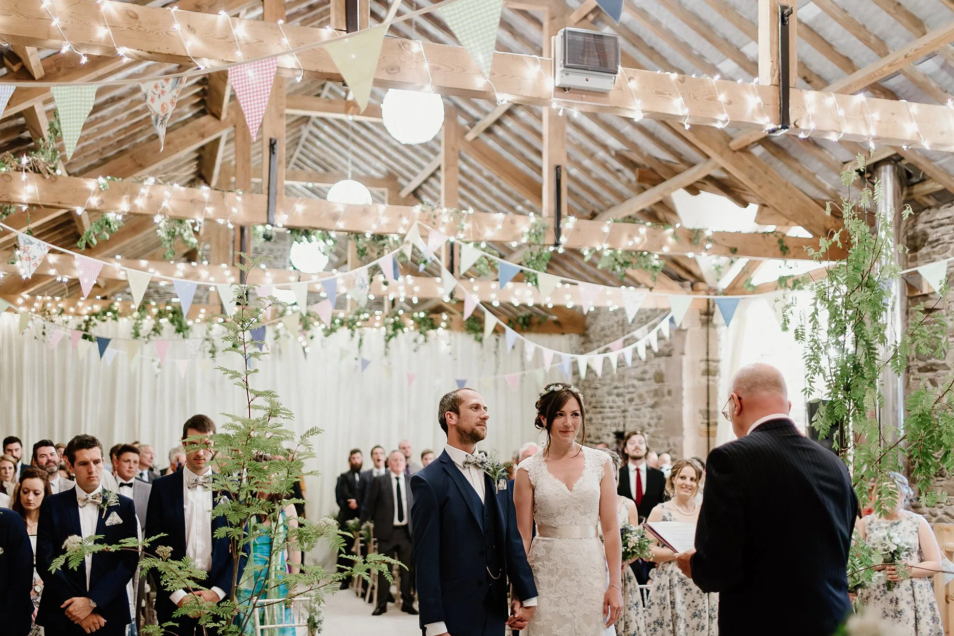 Bride and groom holding hands stood at the front of their wedding ceremony at New House Farm in the Lake District. The ceremony barn is decorated with bunting on the wooden beams.