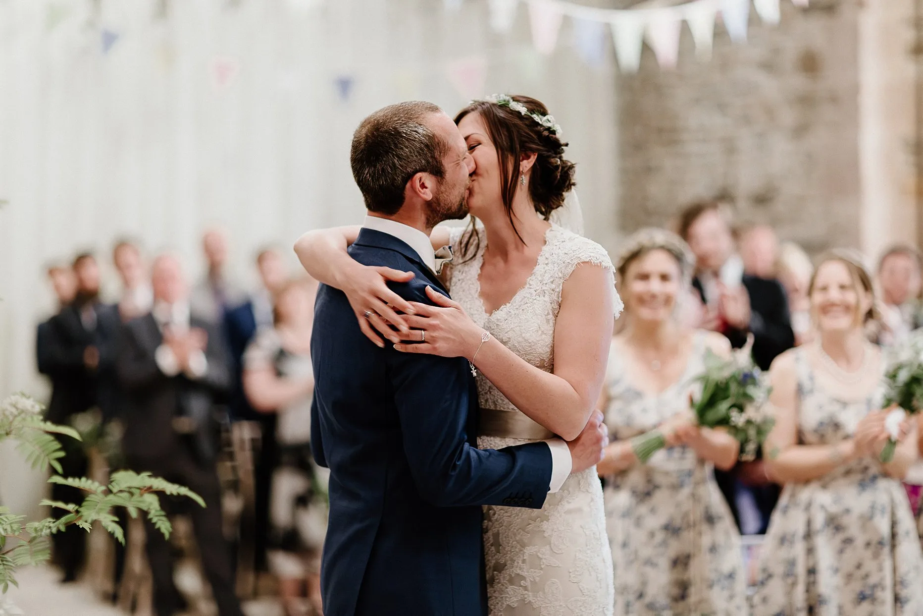 Bride and groom share their first kiss as husband and wife. Guests smiling in the background. The wedding ceremony is in an old barn in the Lake District.