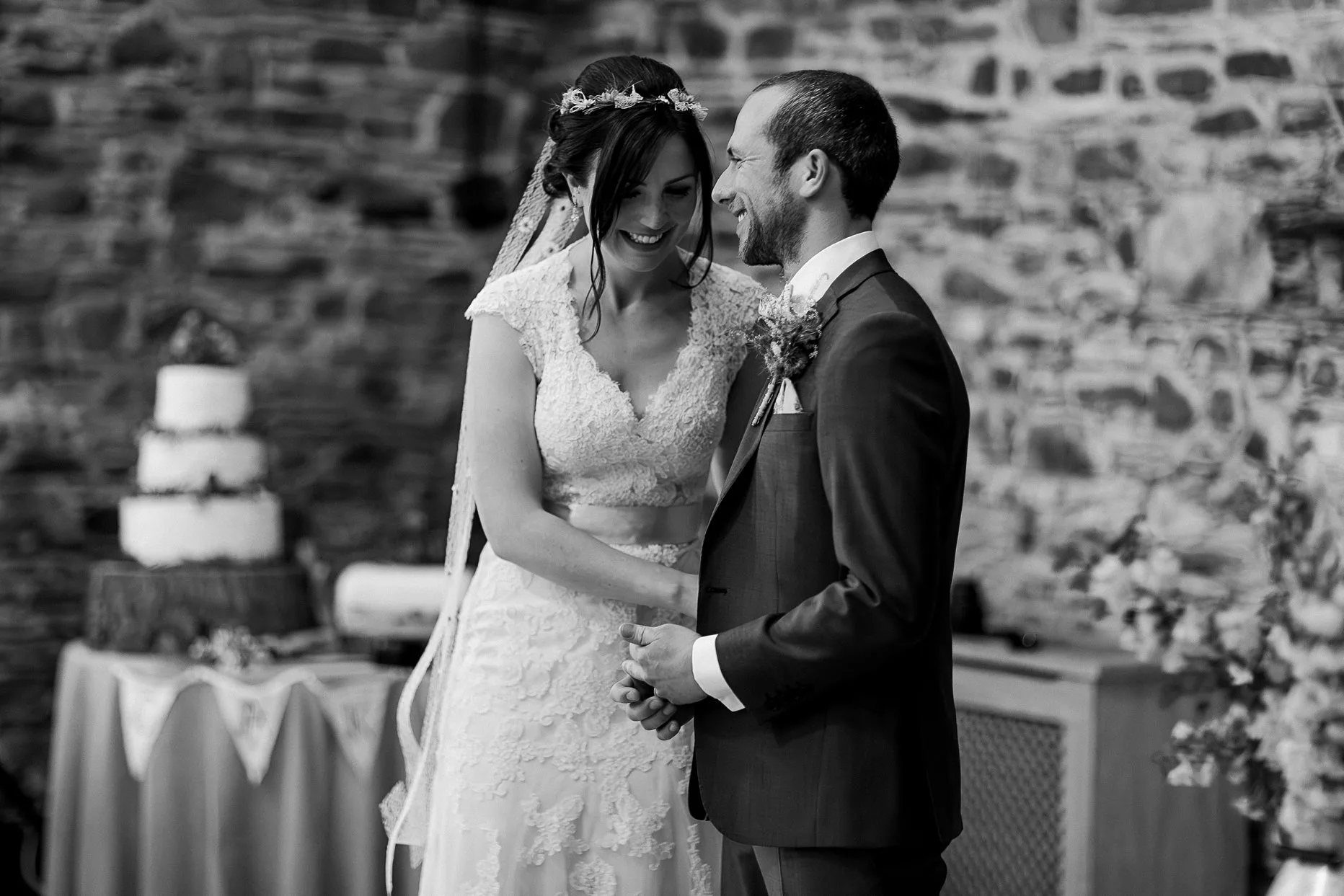 Natural black and white photograph of bride and groom talking and smiling during their wedding ceremony. They are stood in a rustic stone wedding barn.