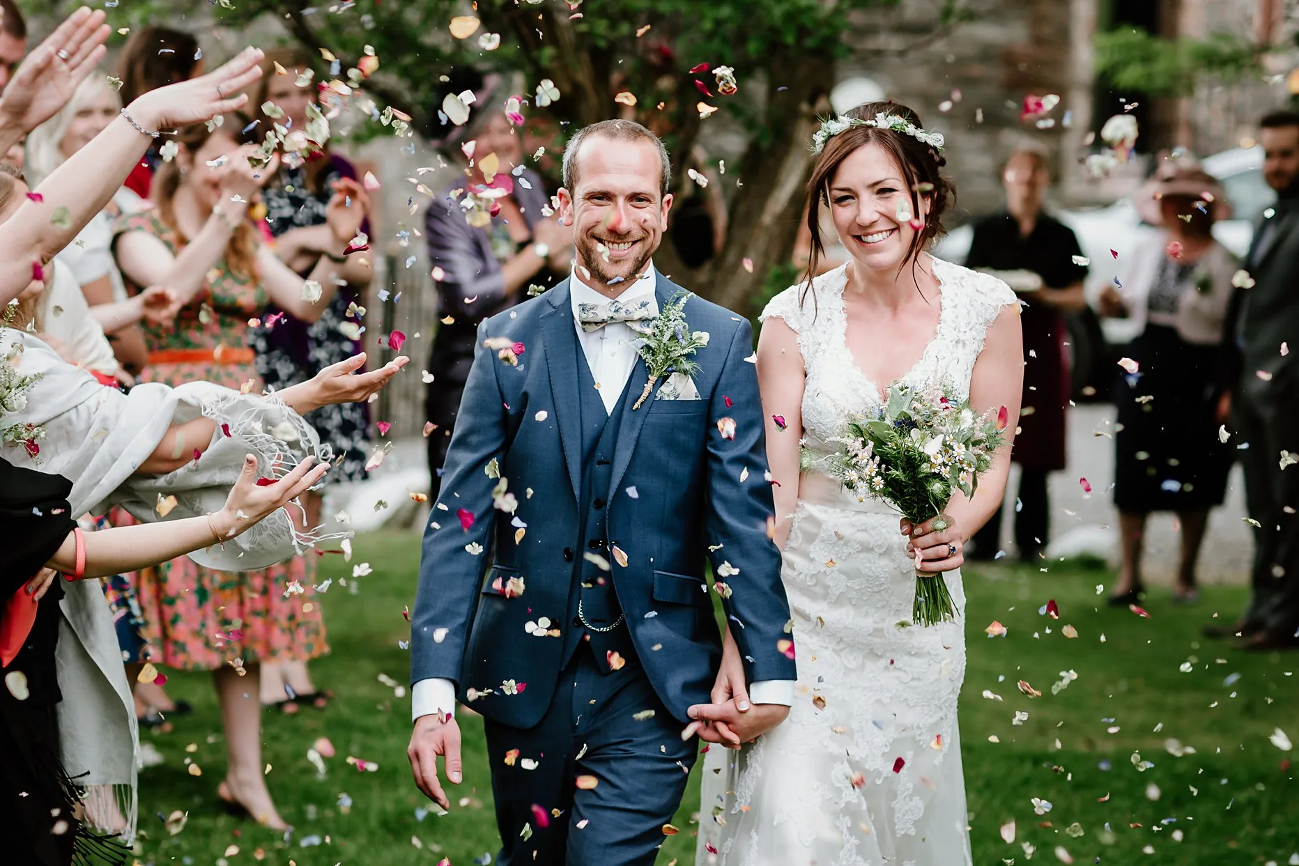 Bride and groom walking through confetti tunnel. Wedding guests stand either side of them throwing confetti. Natural photograph of the bride and groom are smiling and laughing. 