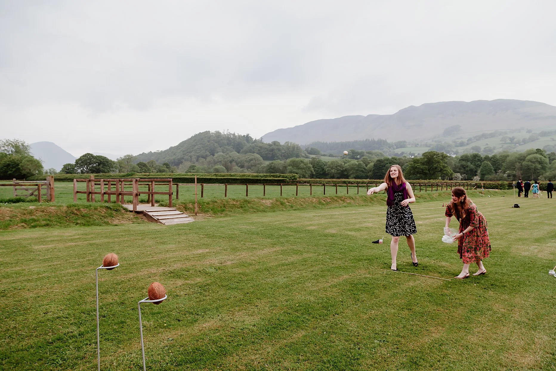 Two female wedding guests playing a game of coconut shy on the garden outside New House Farm wedding barn. In the distance you can see the surrounding hills of the Lake District.