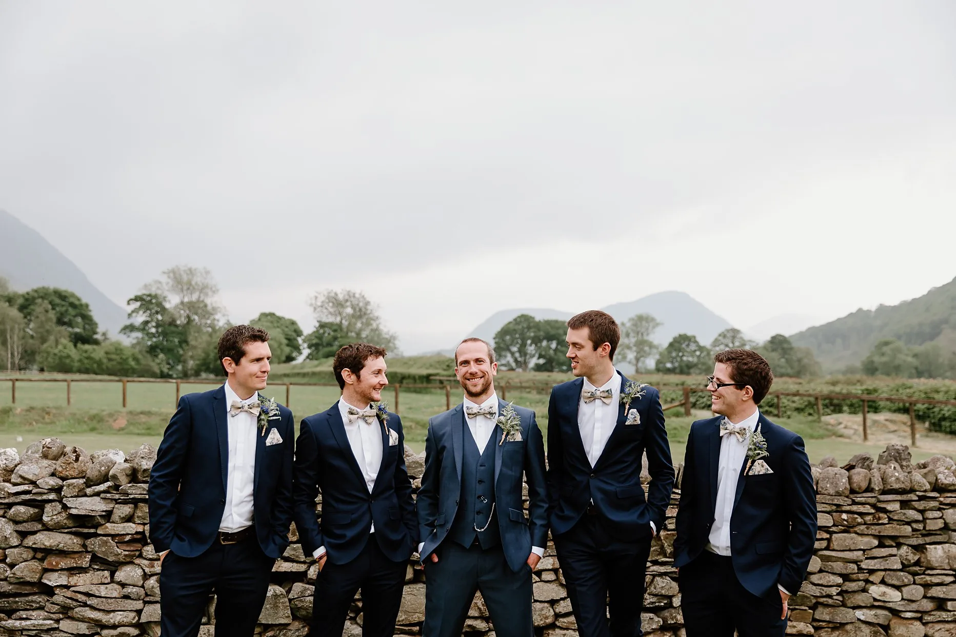 Groom stood with four groomsmen against stone wall in the gardens of New House Farm. All the men are wearing blue suits with a floral bow tie and have their hands in pockets. Groomsmen are looking towards the groom. Beautiful Lake District scenery can be seen in the background.