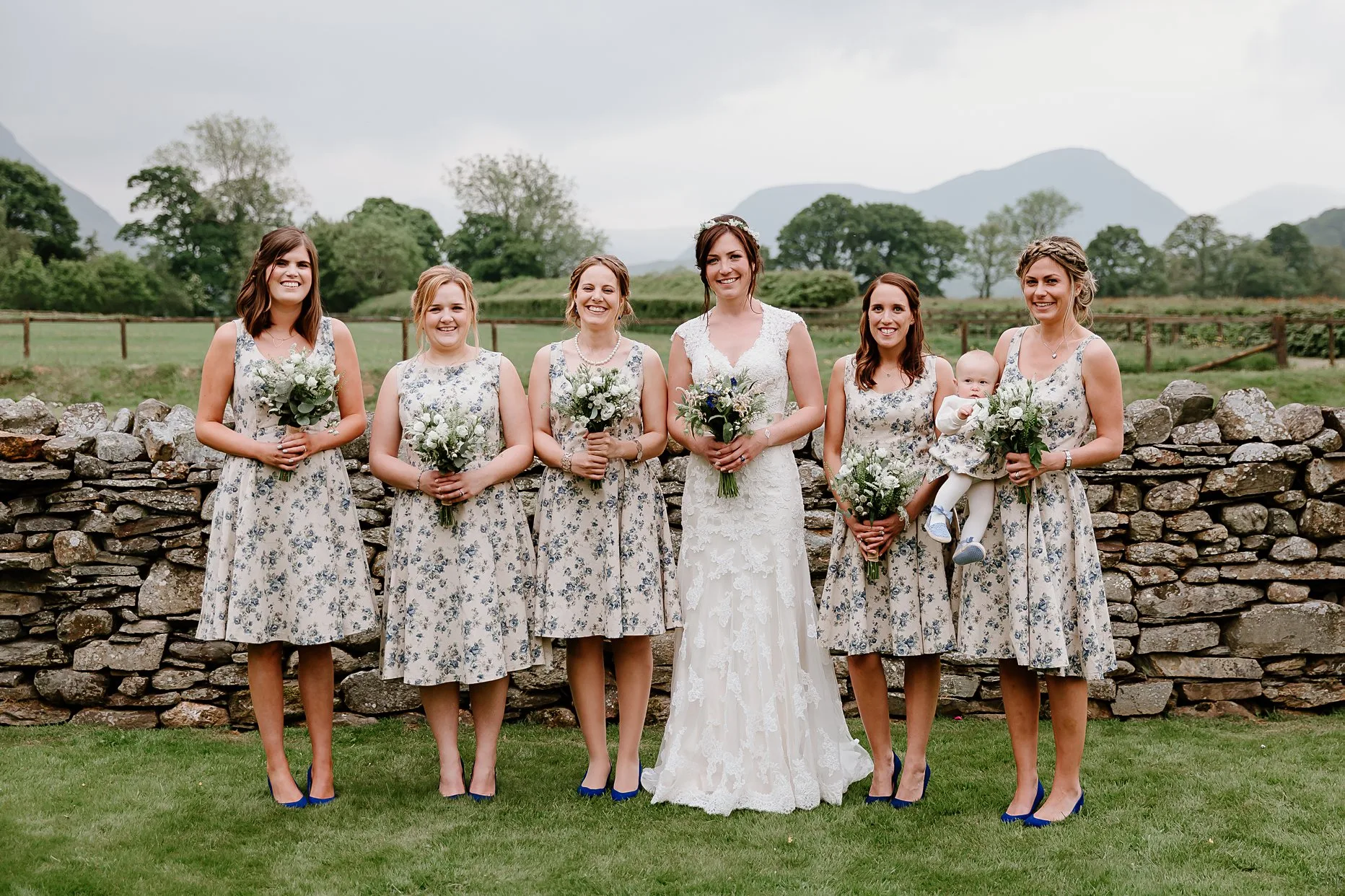 Bride stood with 5 bridesmaids all holding wedding bouquets. Bridesmaids are wearing blue floral dresses and cobalt blue shoes.