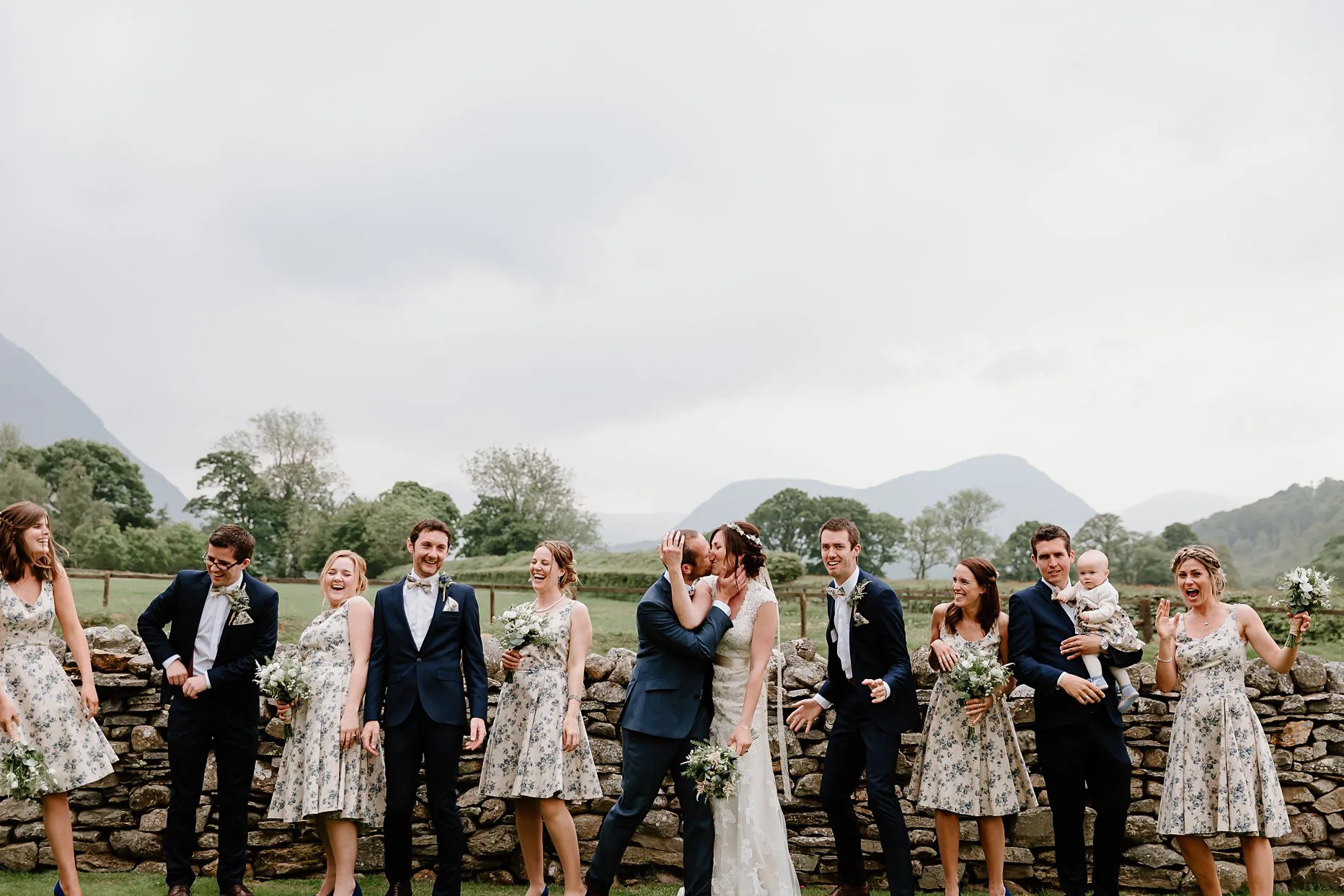 bridesmaids and groomsmen stood in front of a stone wall outside new house farm wedding barn. The wedding party are laughing whilst the bride and groom kiss.