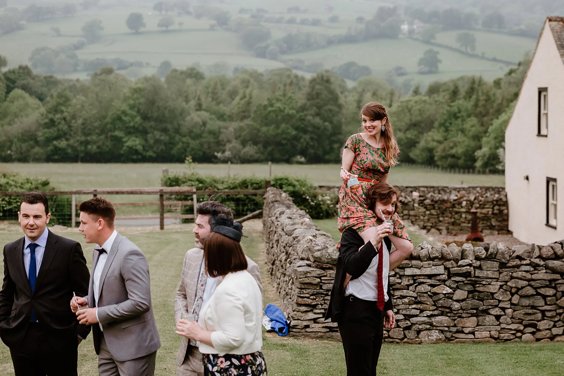 Female wedding guest on male wedding guests shoulders. Both are smiling at the camera with the rolling fields of the Lake District in the background.