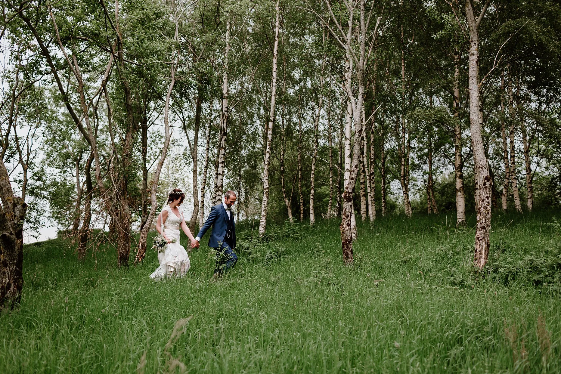 Bride and groom walking through long grass surrounded by silver birch trees in the garden of New House Farm.