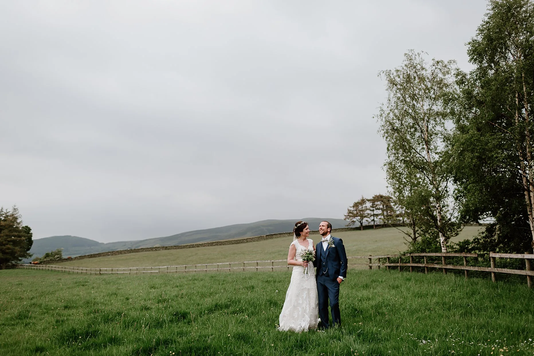 Bride and groom stood smiling and chatting to each other in a field outside New House Farm. Hills and trees in the background. 