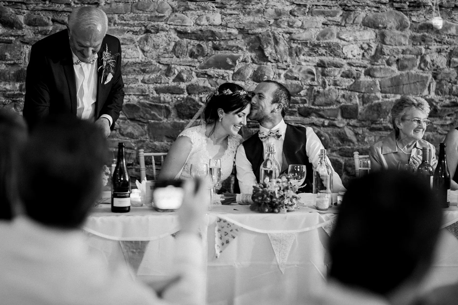 Black and white natural photographer of groom kissing brides forehead during wedding speeches in a wedding barn.