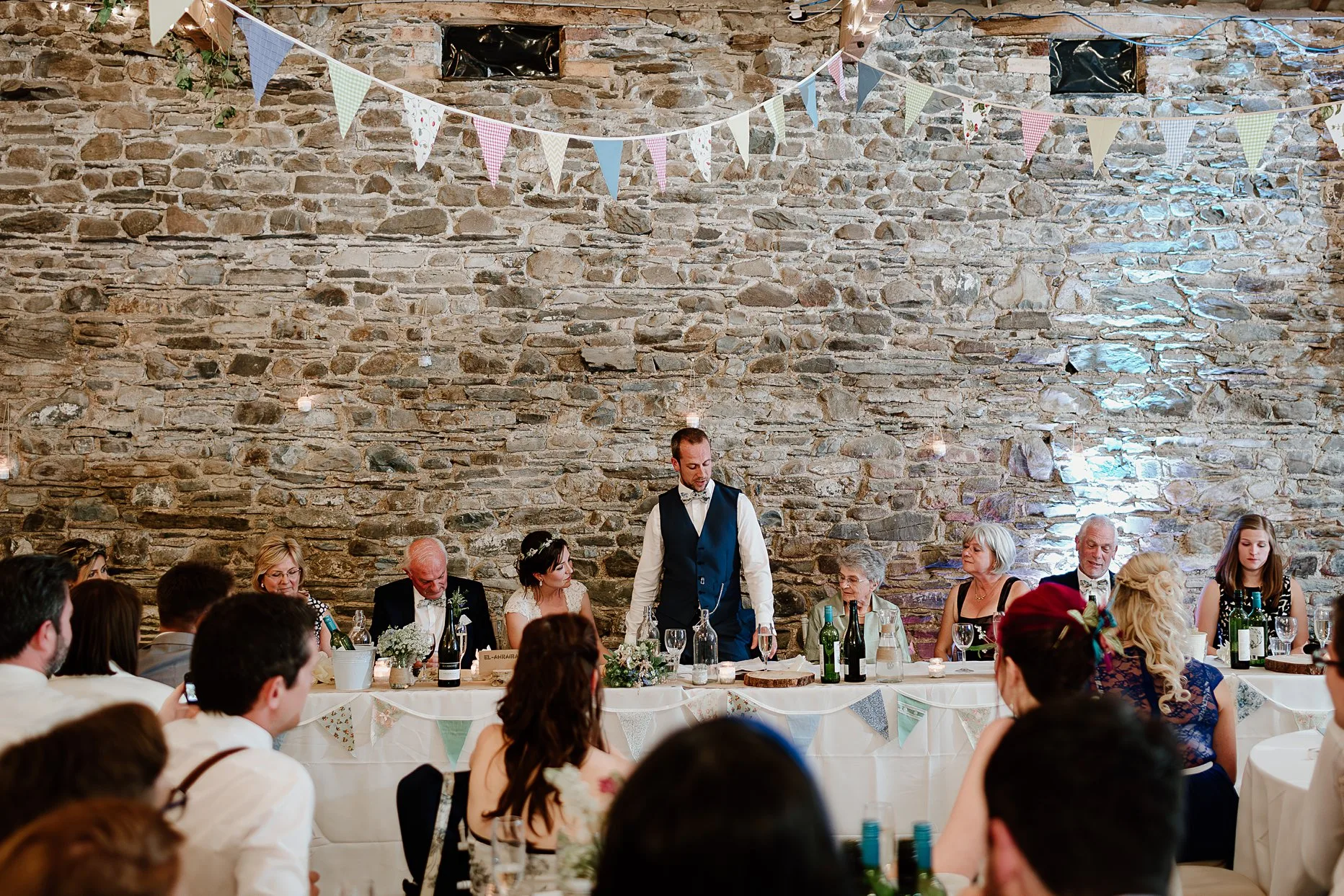 Groom making his wedding speech surrounded by family. He is stood up at a long table in a wedding barn.