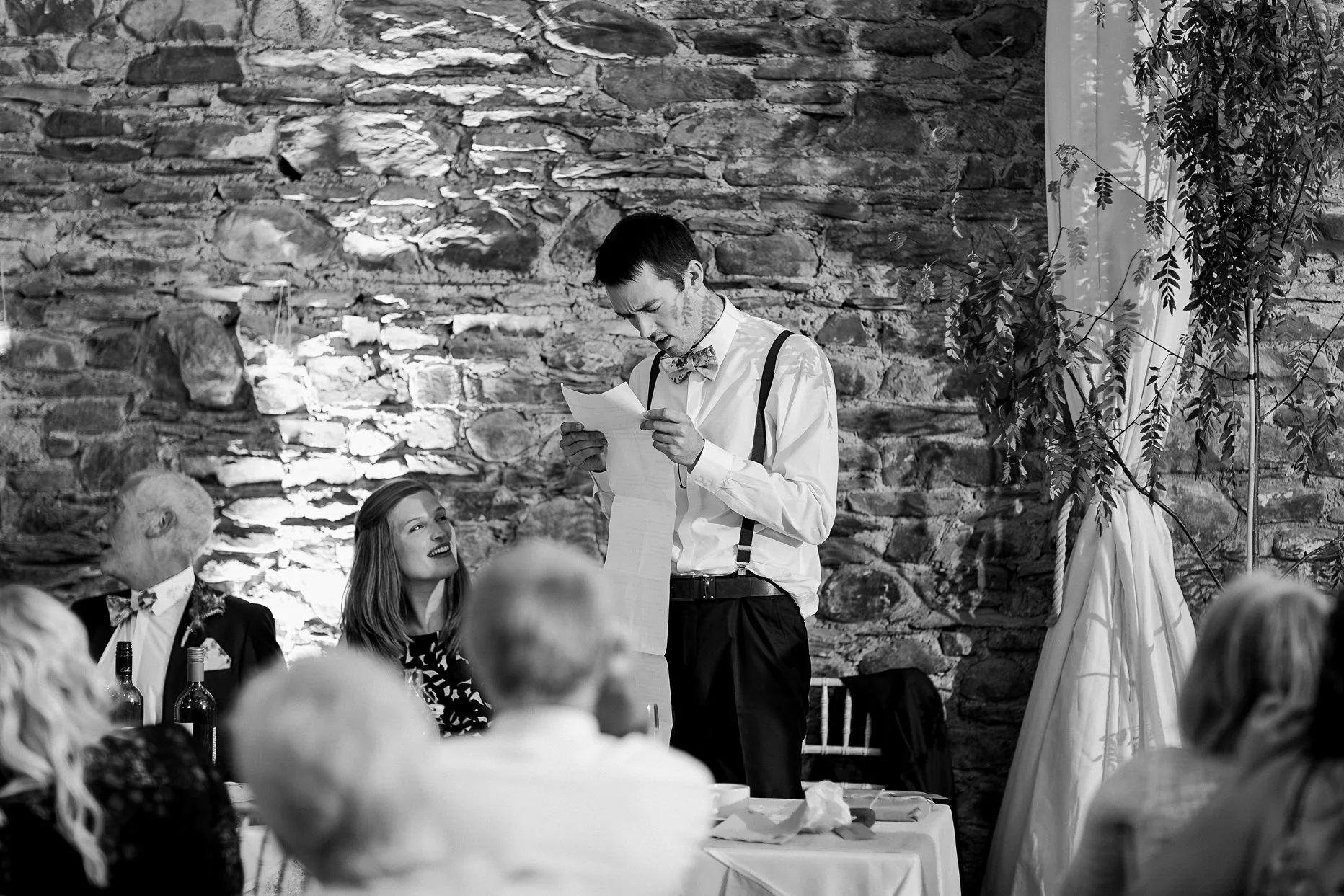 Best man making his wedding speech. He is holding a long sheet of paper and guest around him are laughing.
