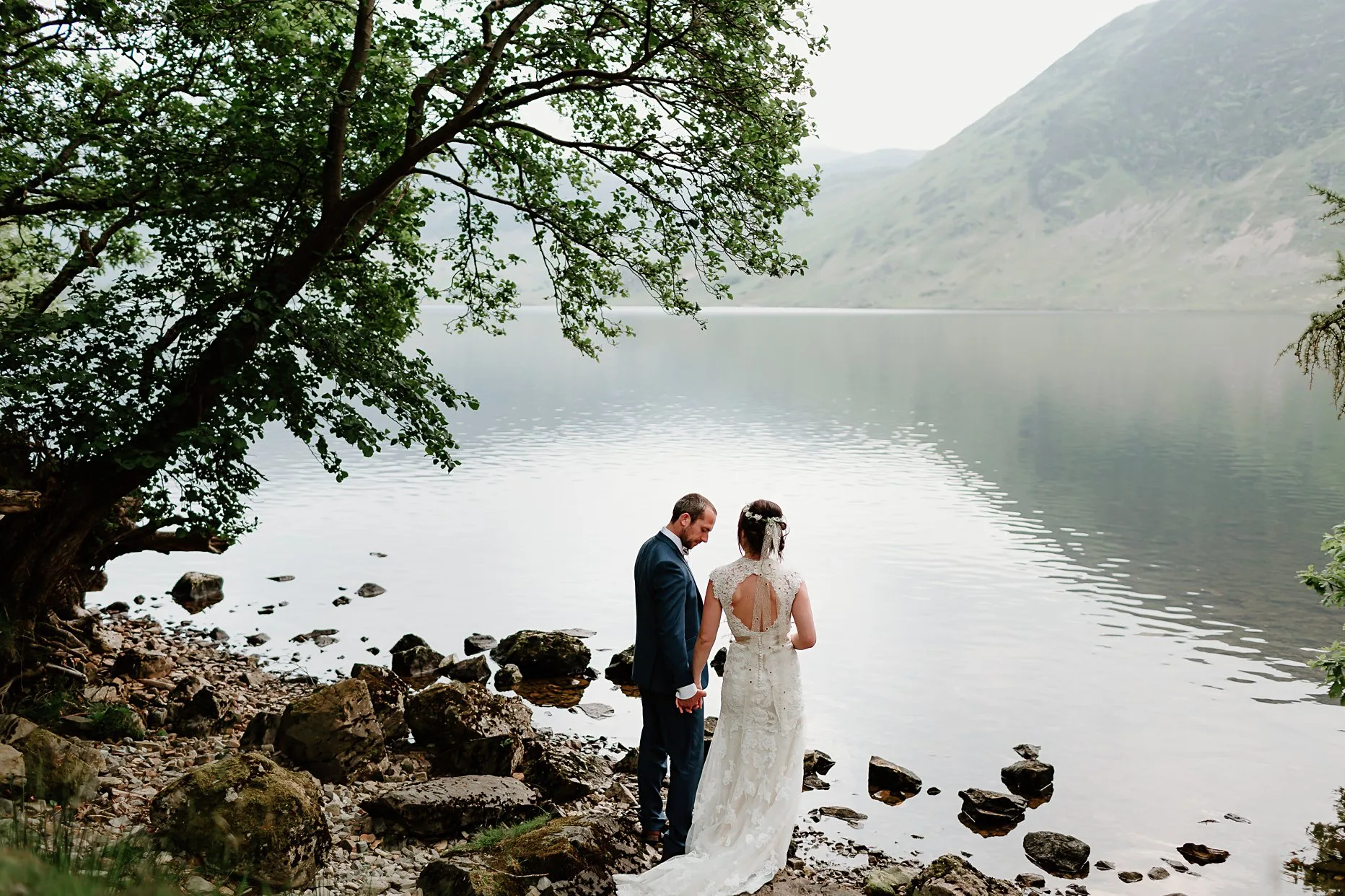 Bride and groom stood facing Crummock Water in the Lake District. The couple are holding hands and groom is looking down to the floor. Bride is wearing a flower crown.