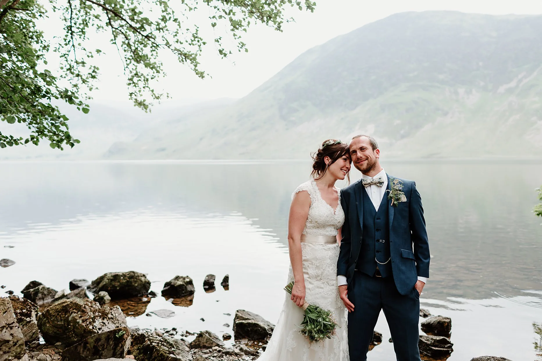 Bride resting head on grooms temple. Groom has his hand in his pocket and both are smiling. Crummock water in the background.